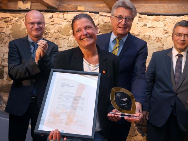 Preisträgerin Agnes Becker hält Ihre Urkunde und den Preis in den Händen und lächelt in die Kamera. Im Hintergrund stehen Fritz Brickwedde, Stiftungsratsvorsitzender und zwei weitere Herren.