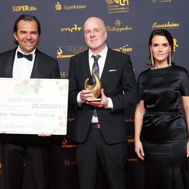 Robert Dahl, Peter Nitschke und Yara Hoffmann stehen vor einer Fotowand mit Trophäe und großem Spendenscheck in der Hand.